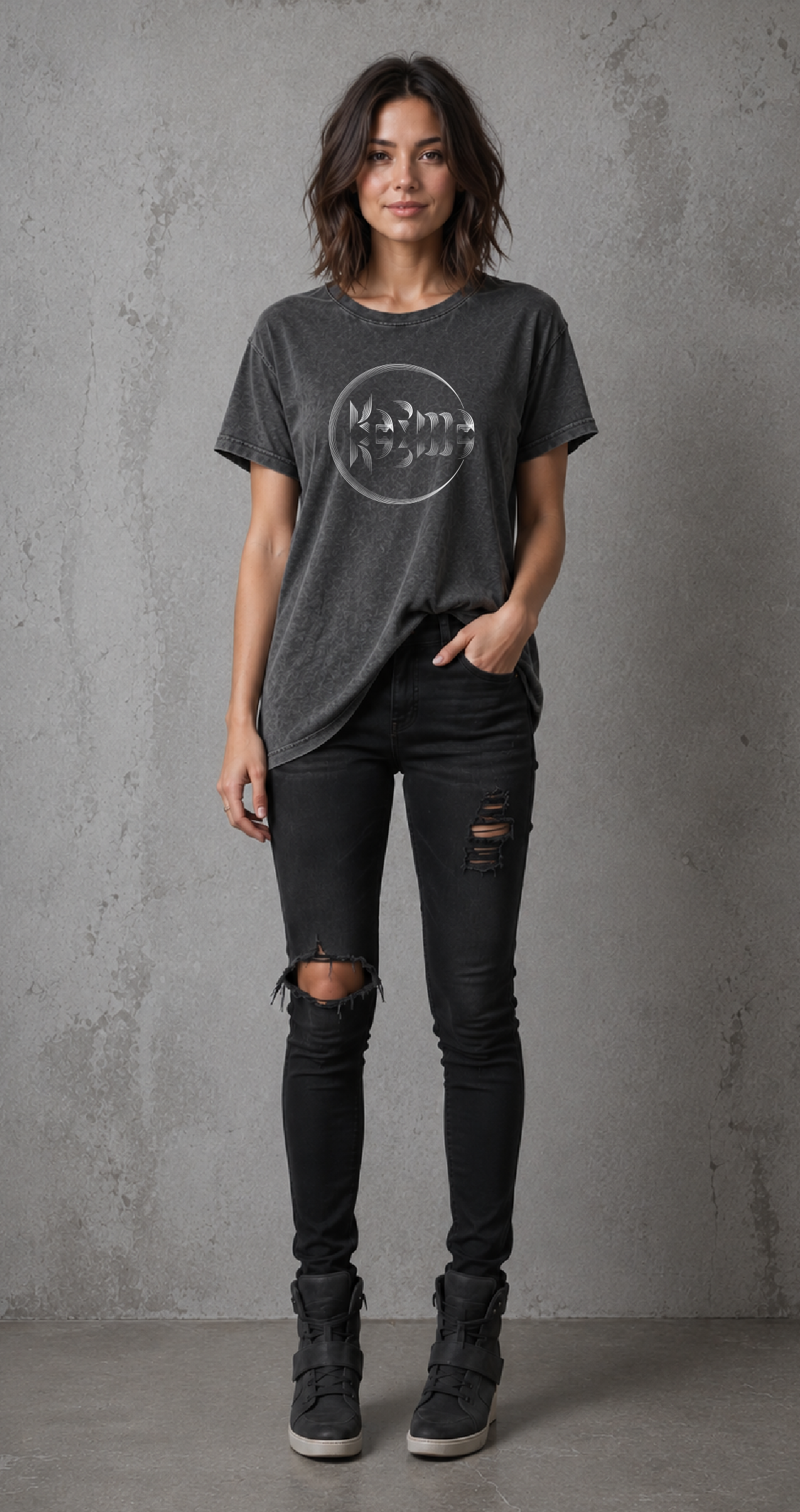 A woman wearing a dark gray washed style t-shirt with a Karma graphic and a ripped black skinny jeans and black lace-up boots, standing against a textured gray concrete wall.