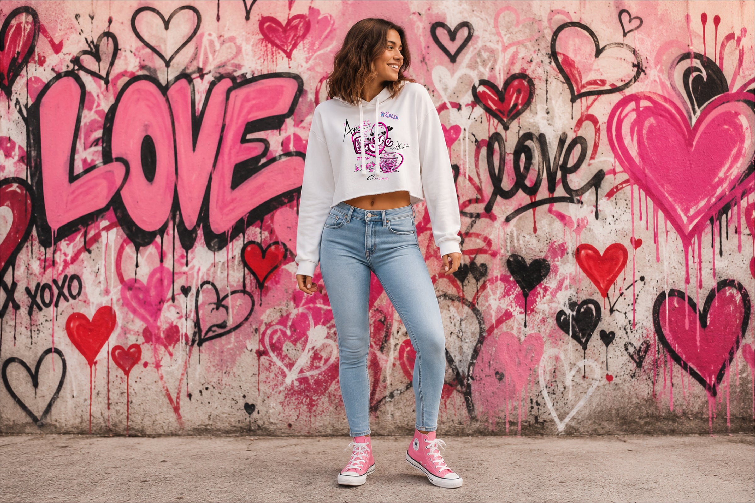 A woman stands in front of a graffiti-covered wall wearing a white cropped hoodie with a original love graphic in different languages, light blue skinny jeans, and pink high-top sneakers. The wall features pink hearts, the word 'LOVE,' and graffiti-style lettering.