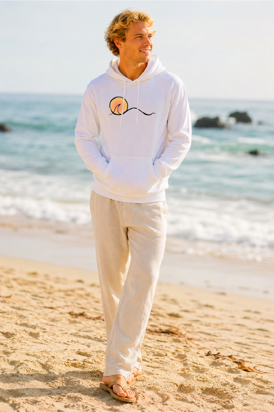 A man standing on a sandy beach wearing a white pullover hoodie with a sunset graphic on the chest, paired with light beige pants and sandals.