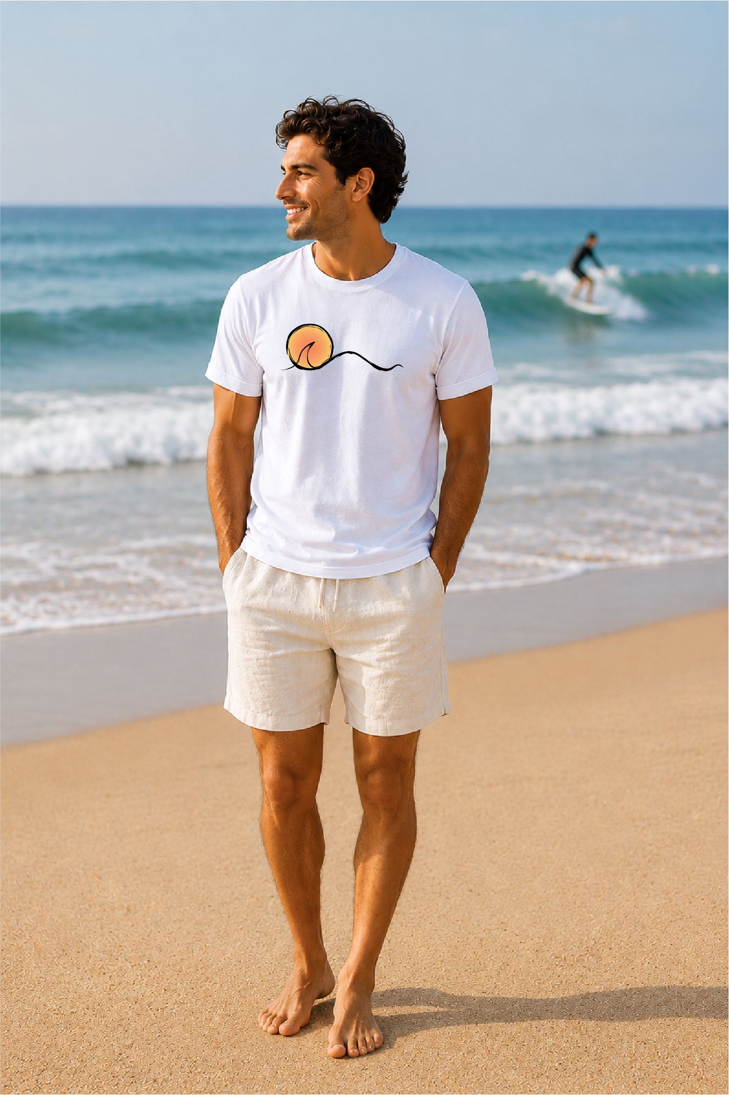 A man standing on a sandy beach wearing a white short-sleeve t-shirt with a sunset graphic on the chest, paired with light beige shorts. The background shows the blue ocean and sky with a surfer in the distance.