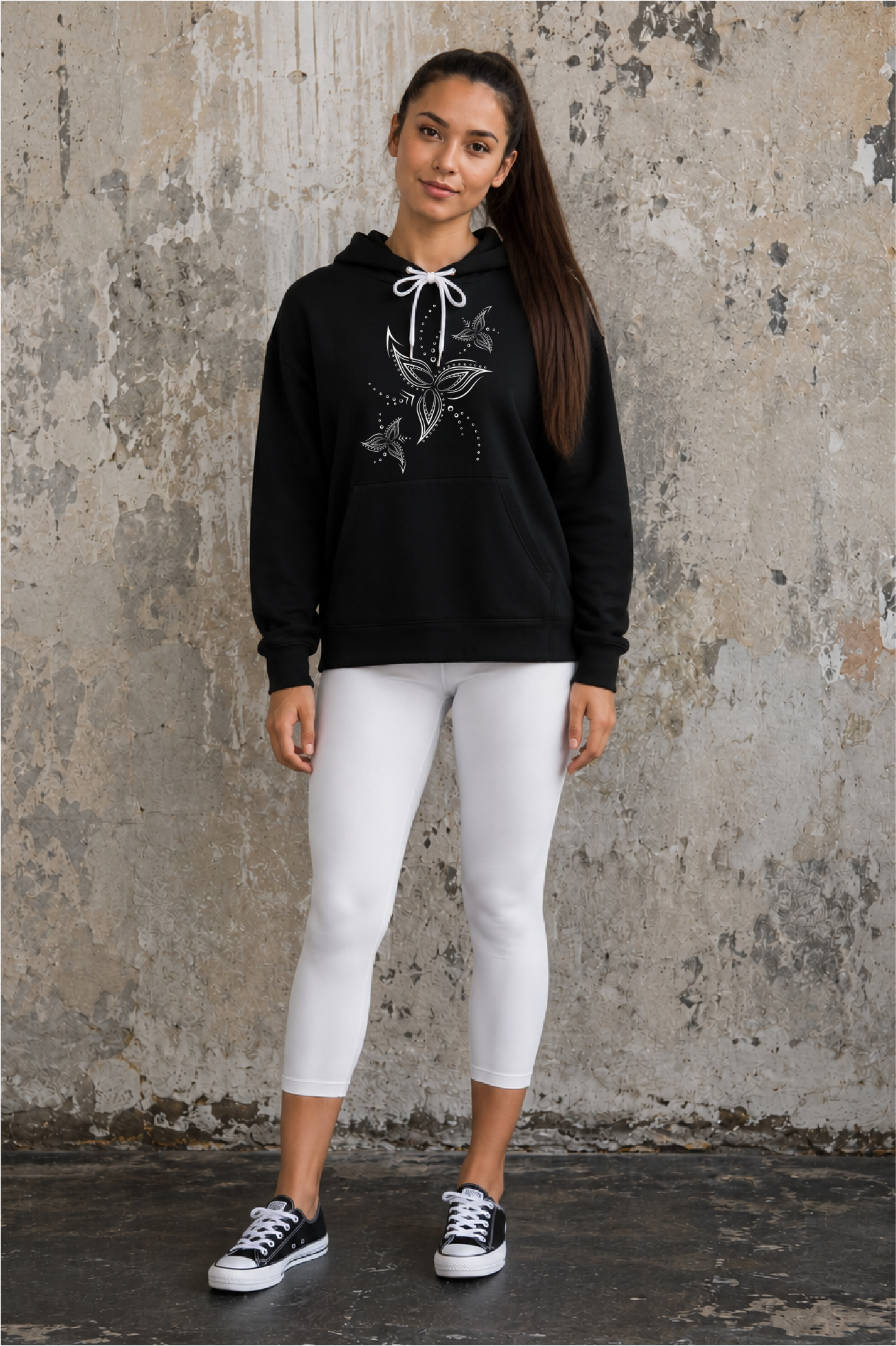A woman stands against a textured concrete wall wearing a black pullover hoodie with a white lined flower graphic, white cropped leggings, and black-and-white sneakers.