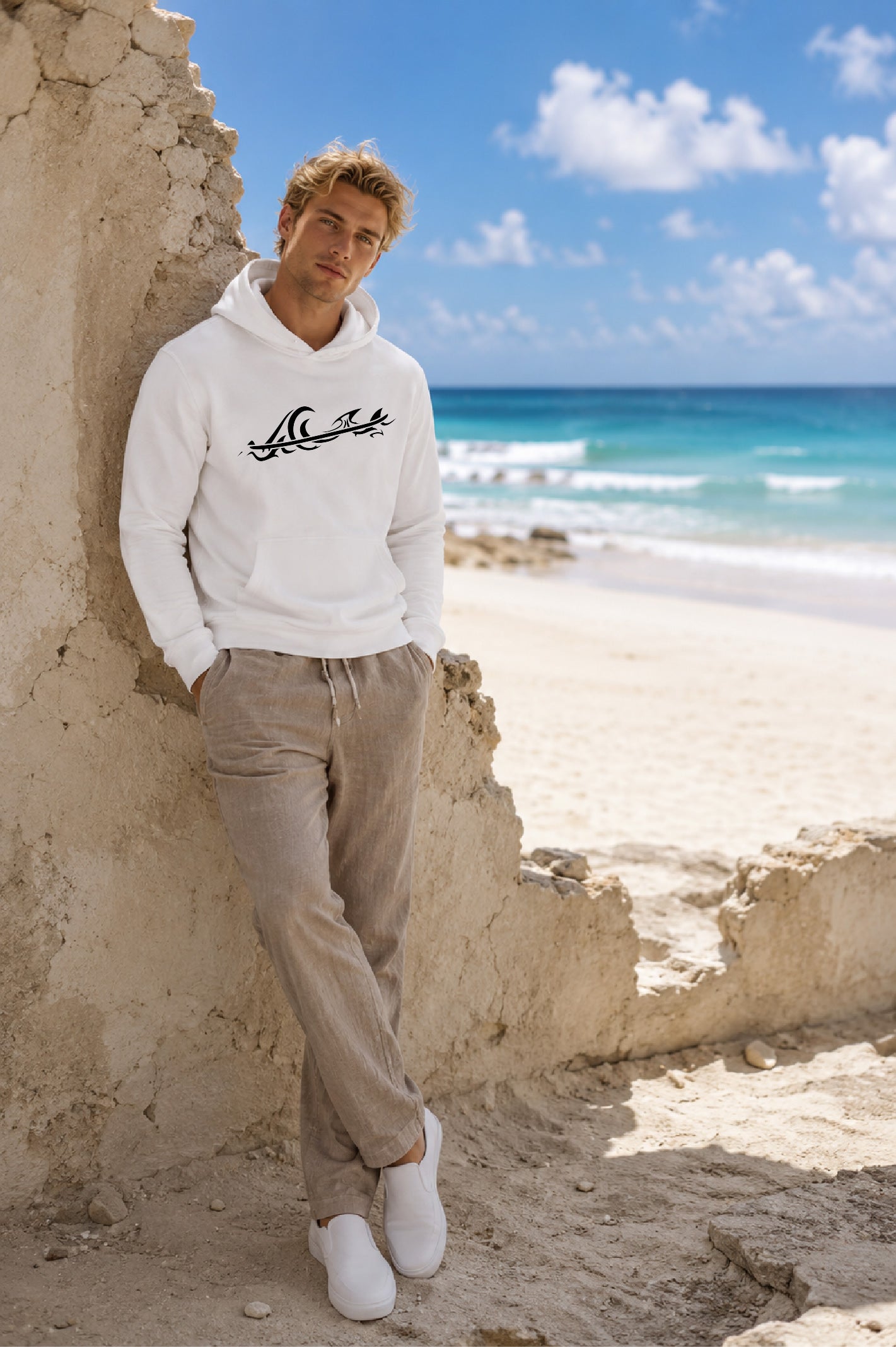 A man stands on a sandy beach leaning against a weathered stone wall, wearing a white hoodie with a black tribal style wave with a surfboard cutting across the wave, beige pants, and white sneakers.