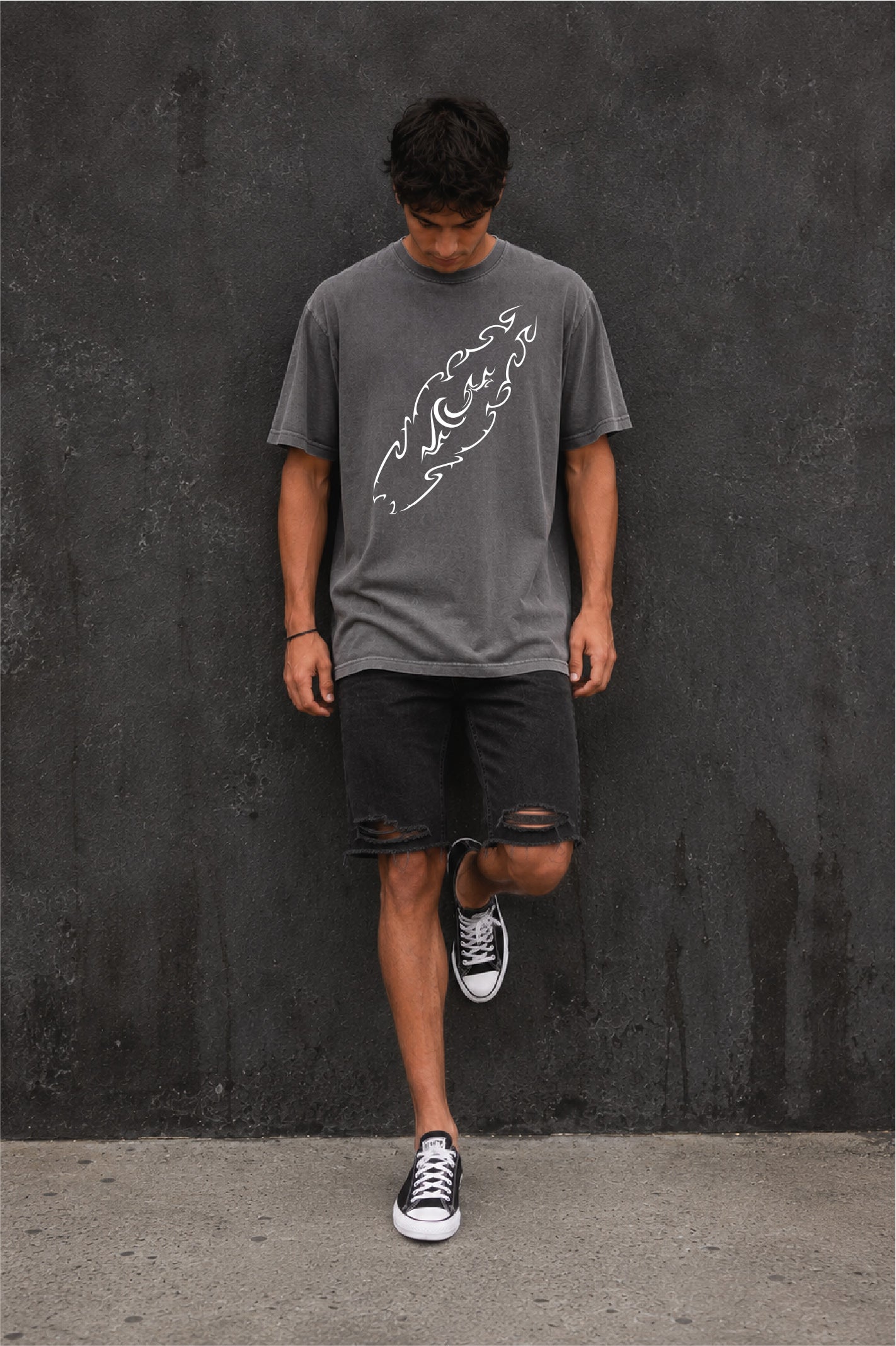 A man wearing a dark gray washed style t-shirt with a white tribal style surfboard graphic with black distressed shorts, and black sneakers, standing against a dark textured wall.