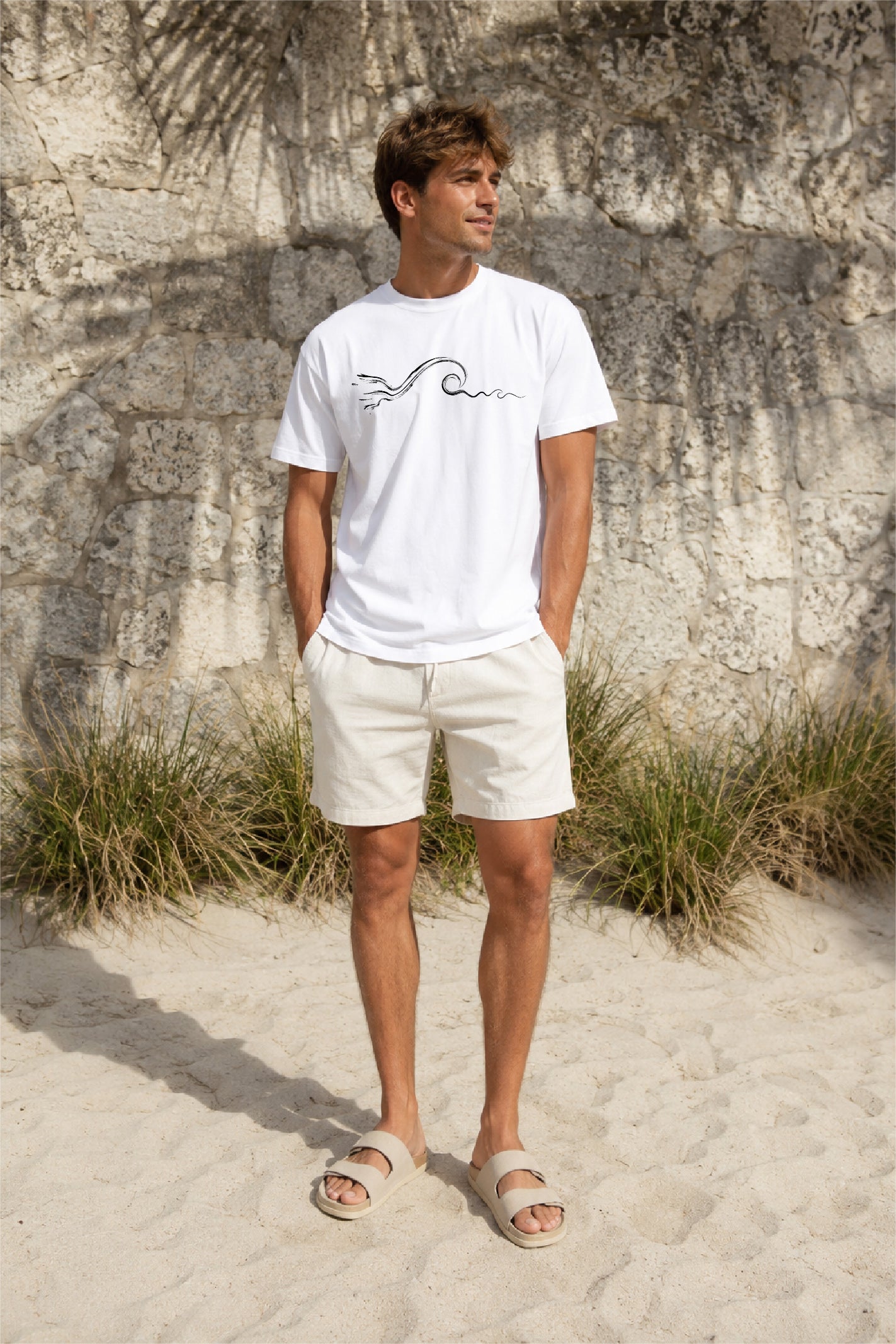 A man standing on a sandy beach in front of a textured rock wall, wearing a white crewneck t-shirt with a black wave graphic, off-white shorts, and beige slide sandals.