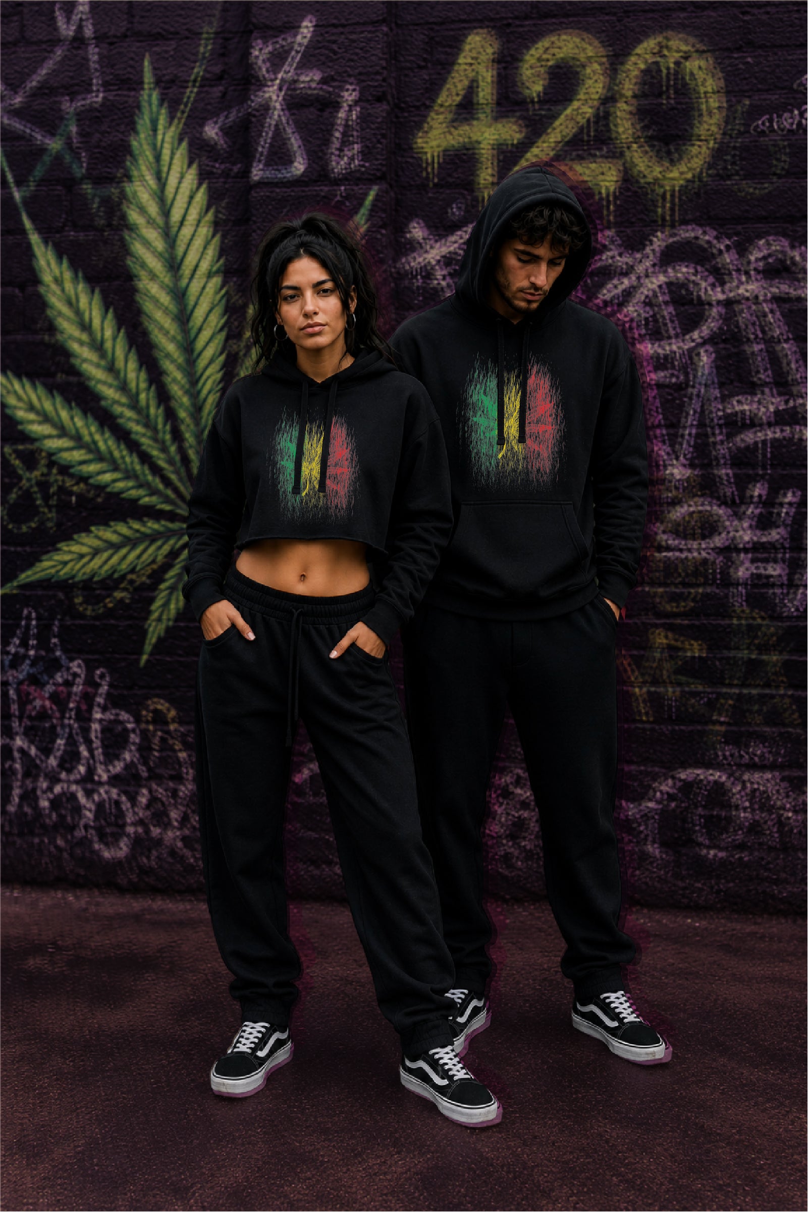 A man and a woman wearing matching black hoodies with a colorful Rastafarian rain effect color with a hemp leaf graphic, standing in front of a graffiti-covered wall.