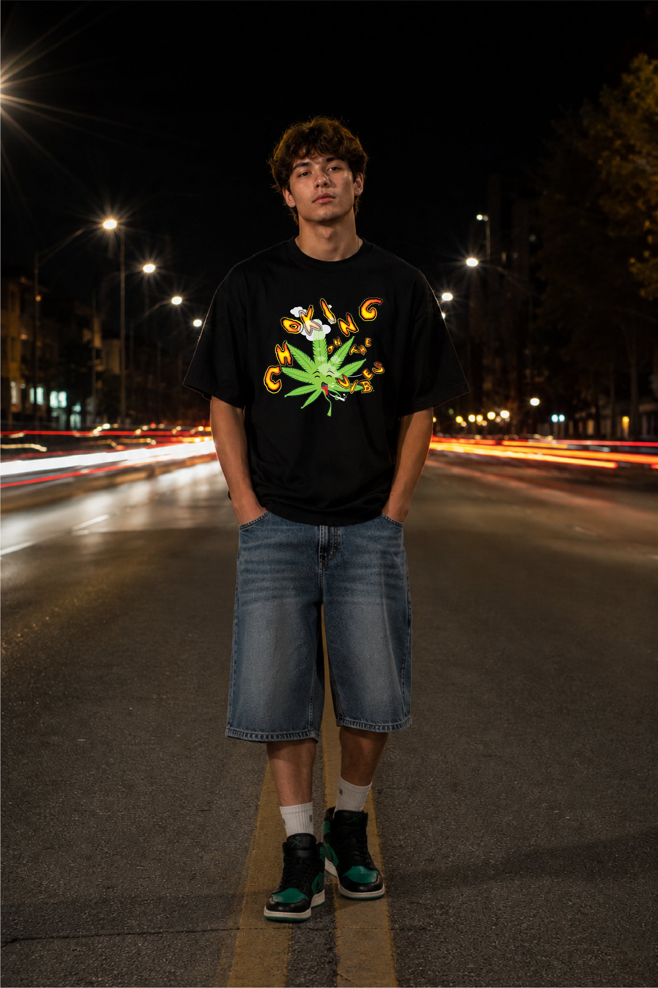 A young man stands on a city street at night wearing a black graphic t-shirt with a funny choking cannabis leaf design, blue denim shorts, white socks, and dark sneakers.
